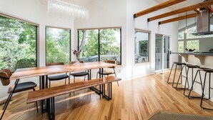 A modern dining area with a wooden table and benches, large windows, and a kitchen area with bar stools. - Treetop Castle *Sleeps 16* (Boise)