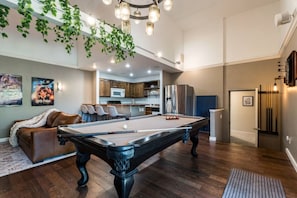 A pool table in a living room with a sofa and a kitchen in the background. - Treetop Castle *Sleeps 16* (Boise)