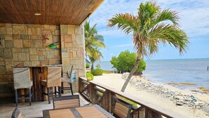 Een terras met stenen vloer en houten meubilair, met uitzicht op een strand met palmbomen en een heldere lucht.