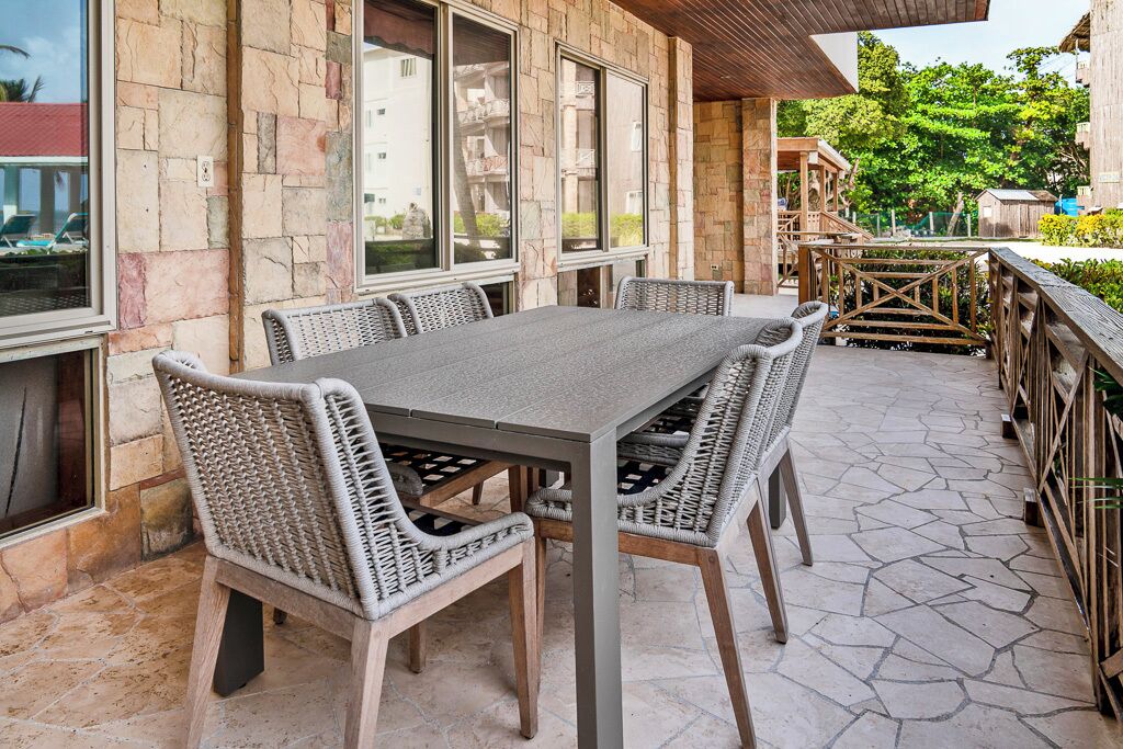 Un patio de piedra con una mesa rectangular y sillas de mimbre, rodeado de muros de piedra y ventanas amplias.