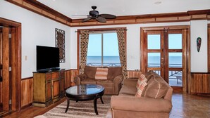 Ein Wohnzimmer mit Fernseher, Sofas, einem Couchtisch und Blick auf den Ozean durch große Fenster.