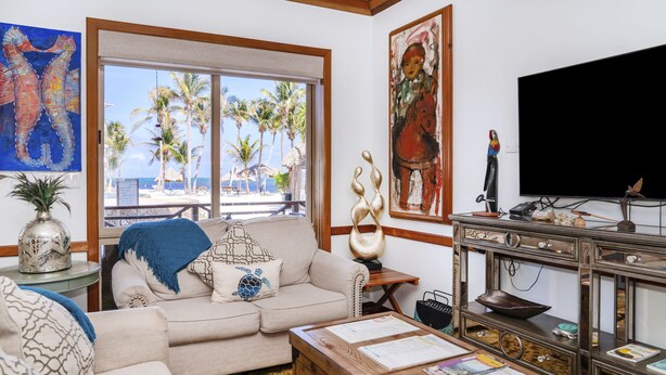 A living room with a beige sofa, a wooden coffee table with papers, a TV, and a view of the beach with palm trees. - Luxury Beachfront Condo - Condo 17 at Oceans 23, Belize (San Pedro)