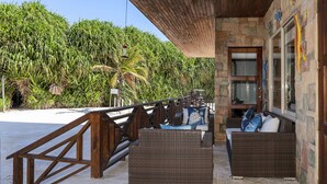 A covered outdoor seating area with wicker furniture, a stone wall, and a view of a beach and palm trees. - Luxury Beachfront Condo - Condo 17 at Oceans 23, Belize (San Pedro)