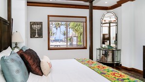 A bedroom with a bed, pillows, a window with a view of palm trees and a beach, a mirror, and a side table. - Luxury Beachfront Condo - Condo 17 at Oceans 23, Belize (San Pedro)
