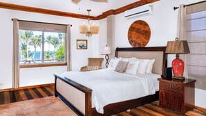 Une chambre avec un lit en bois, une table de chevet, un lustre, une grande fenêtre donnant sur des palmiers et un élément décoratif circulaire sur le mur.
