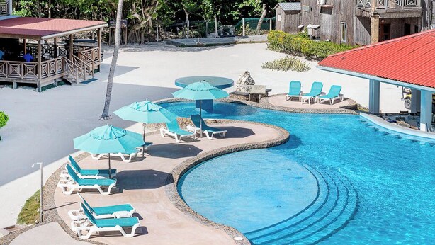 A swimming pool with multiple sun loungers and umbrellas, surrounded by a wooden deck and a building with a red roof.