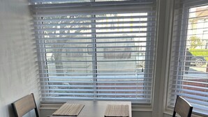 Uma área de refeições com mesa e quatro cadeiras, janelas amplas com persianas e vista para o exterior.