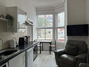 A compact kitchen with a dining table and a leather sofa, a flat-screen TV mounted on the wall, and large windows with a view of the street.