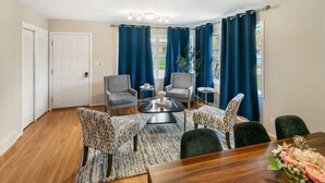 Una sala de estar moderna con suelo de madera, una mesa de comedor con cuatro sillas, dos sillones, una mesita de centro y un ventanal con cortinas azules.