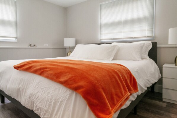 A bedroom with a bed, white bedding, an orange throw blanket, a nightstand, and a lamp. - Newly remodeled 4 bedroom home in DT Libertyville Near Naval Base! (Libertyville)