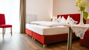 A hotel room with a red bed, white linens, a red chair, a white vase with flowers, and a wooden floor. - Pension Tannenhof (Laces)
