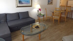 Un salon avec un canapé gris, une table basse en verre, une lampe et un tableau représentant une scène de plage.