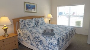 Une chambre à coucher comprenant un lit, deux lampes de chevet, une table de nuit, une fenêtre avec des stores et un tableau encadré accroché au mur.