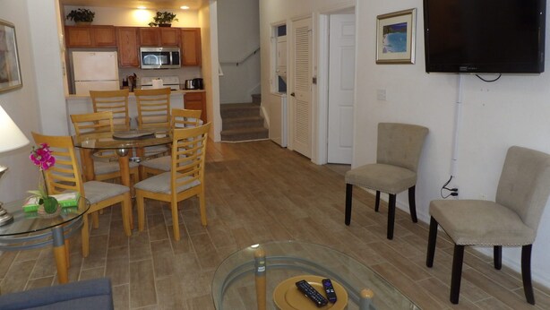 Un salon avec une table basse en verre, un téléviseur fixé au mur et un coin repas comprenant des chaises et une table en bois.