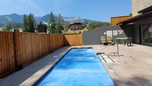 A rectangular swimming pool with a wooden fence and a view of mountains in the background. - Griz Inn Condo on the Hill - 100 steps from lift (Fernie)