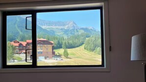 A room with a large window offering a view of a mountain landscape, a building with red roofs, and a parked vehicle. - Griz Inn Condo on the Hill - 100 steps from lift (Fernie)