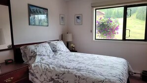A bedroom with a bed, a dresser, a window with a view of greenery, and framed pictures on the wall. - Griz Inn Condo on the Hill - 100 steps from lift (Fernie)