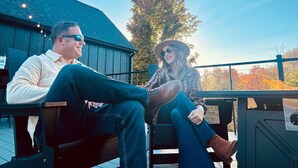 Two people sitting on a wooden deck with a metal railing, a wooden building in the background, and a tree with autumn leaves. - Spillway Lodges (Bellaire)