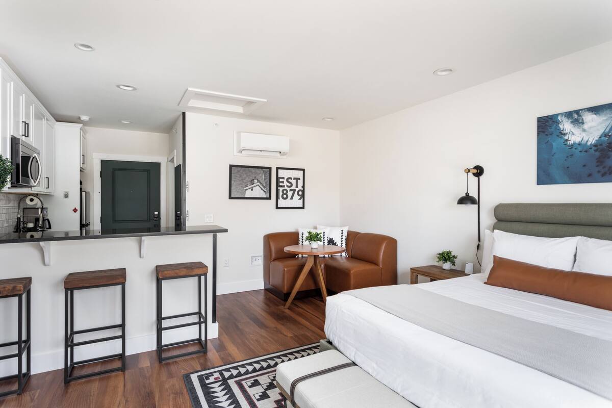 A modern hotel room with a kitchen area, a bed with a large window, and a small seating area.