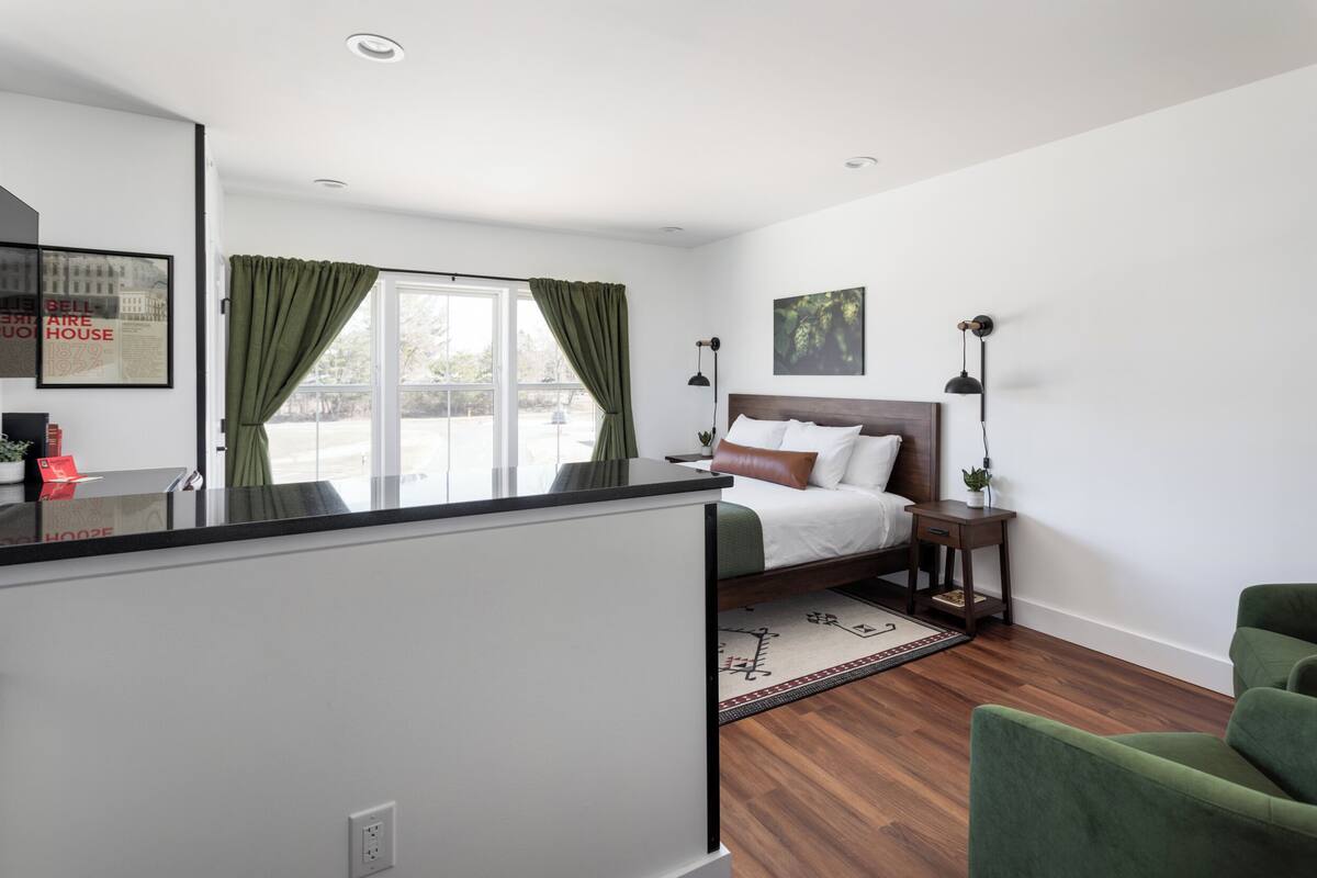 A modern bedroom with a bed, a nightstand, a green chair, and a large window with curtains.