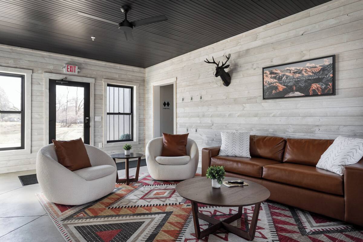 A modern living room with a white and brown leather sofa, two white armchairs, a patterned rug, and a deer head mounted on the wall.