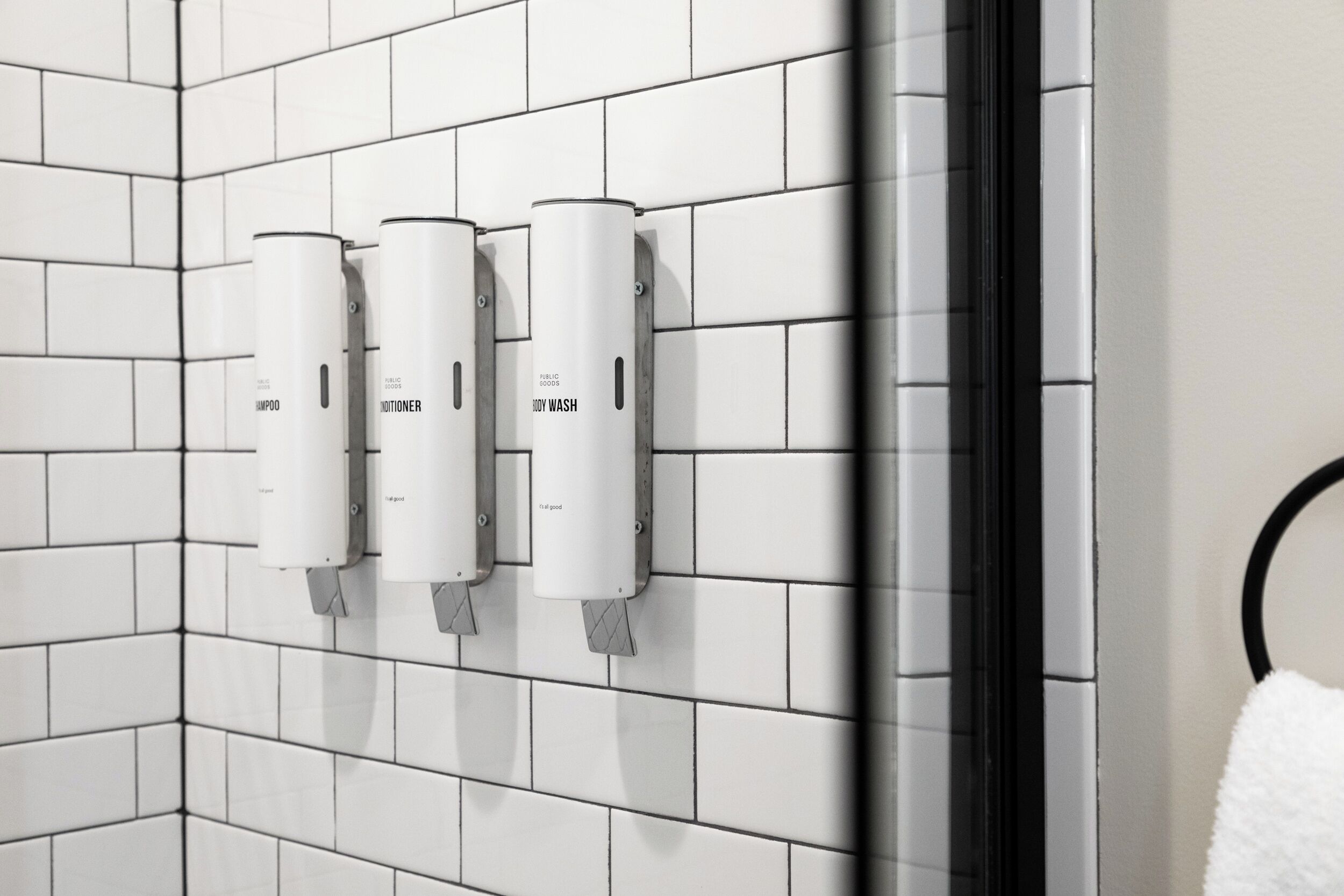 A bathroom with white tiled walls and three dispensers labeled "shampoo," "conditioner," and "body wash."