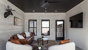 A modern living room with a leather sofa, two white armchairs, a coffee table, and a mounted deer head. There are three windows, a ceiling fan, and a TV mounted on the wall. - Spillway Lodges (Bellaire)