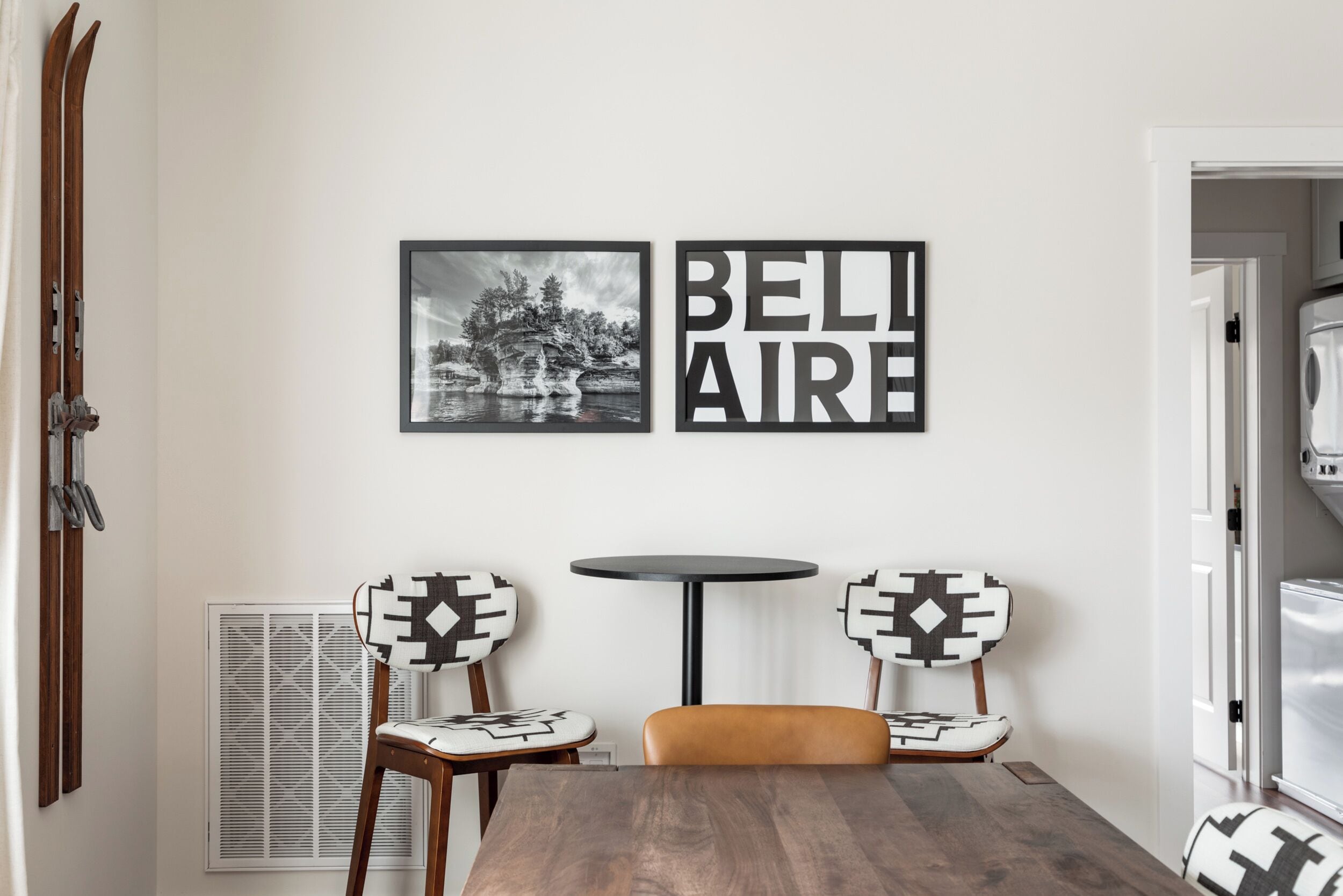 Un comedor con una mesa de madera, dos sillas con cojines estampados, una mesita redonda y dos cuadros en la pared.