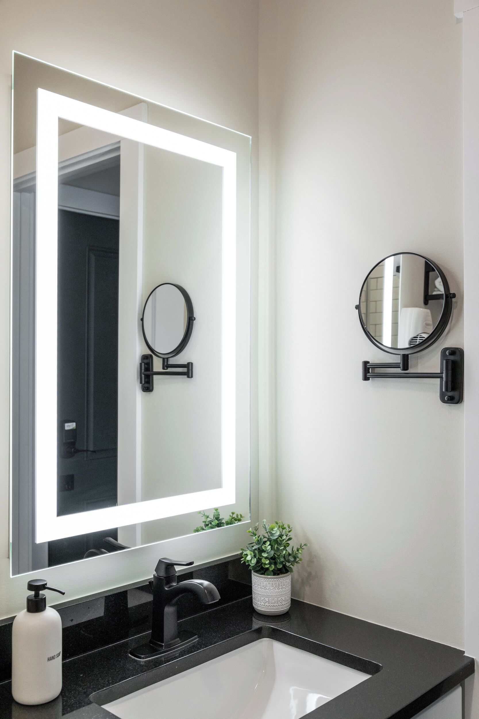 A bathroom with a large mirror, a smaller wall-mounted mirror, a black faucet, a white sink, and a potted plant.