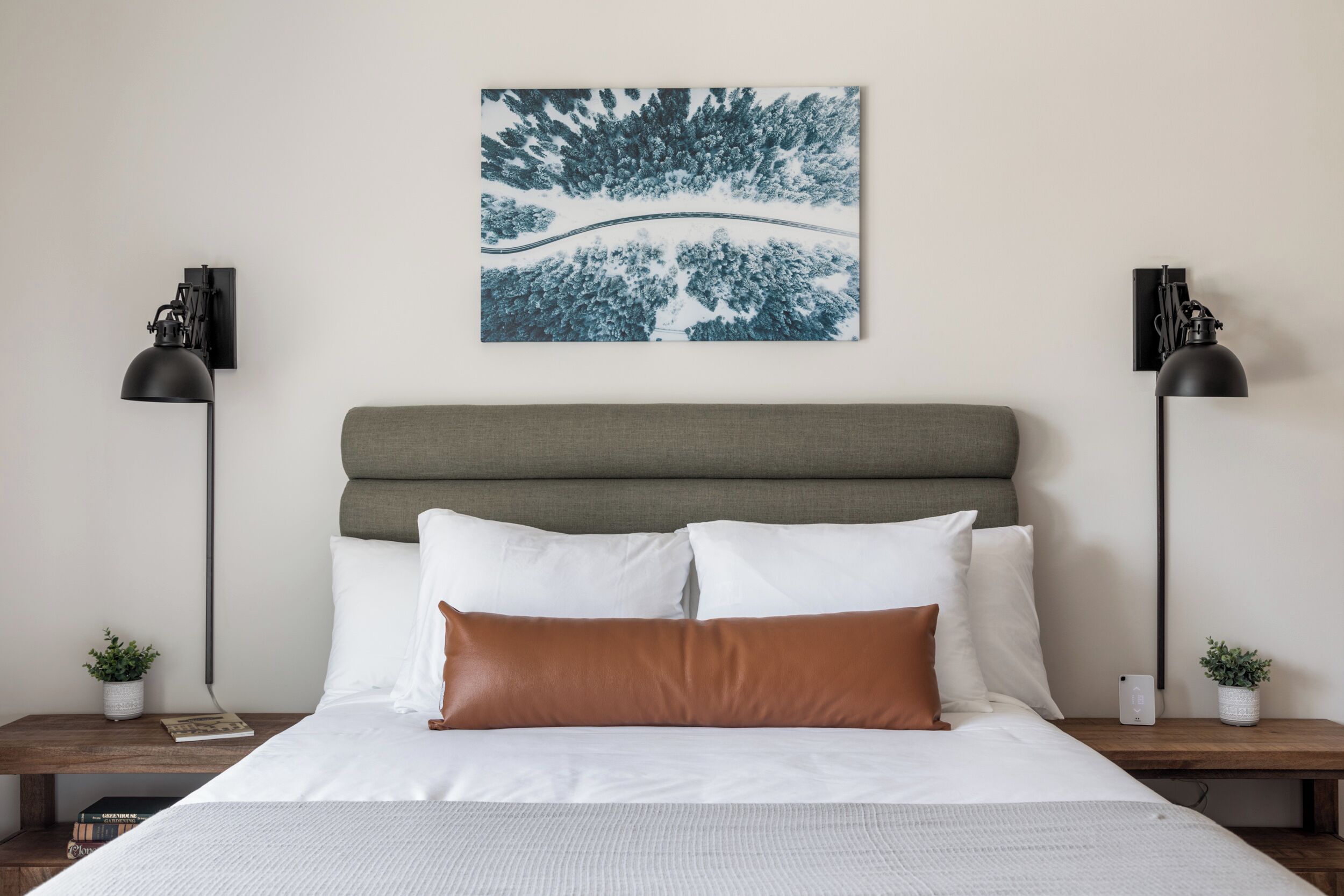 Un lit bien fait, avec une literie blanche et marron, une tête de lit rembourrée de couleur grise et une œuvre d’art abstraite accrochée au mur.