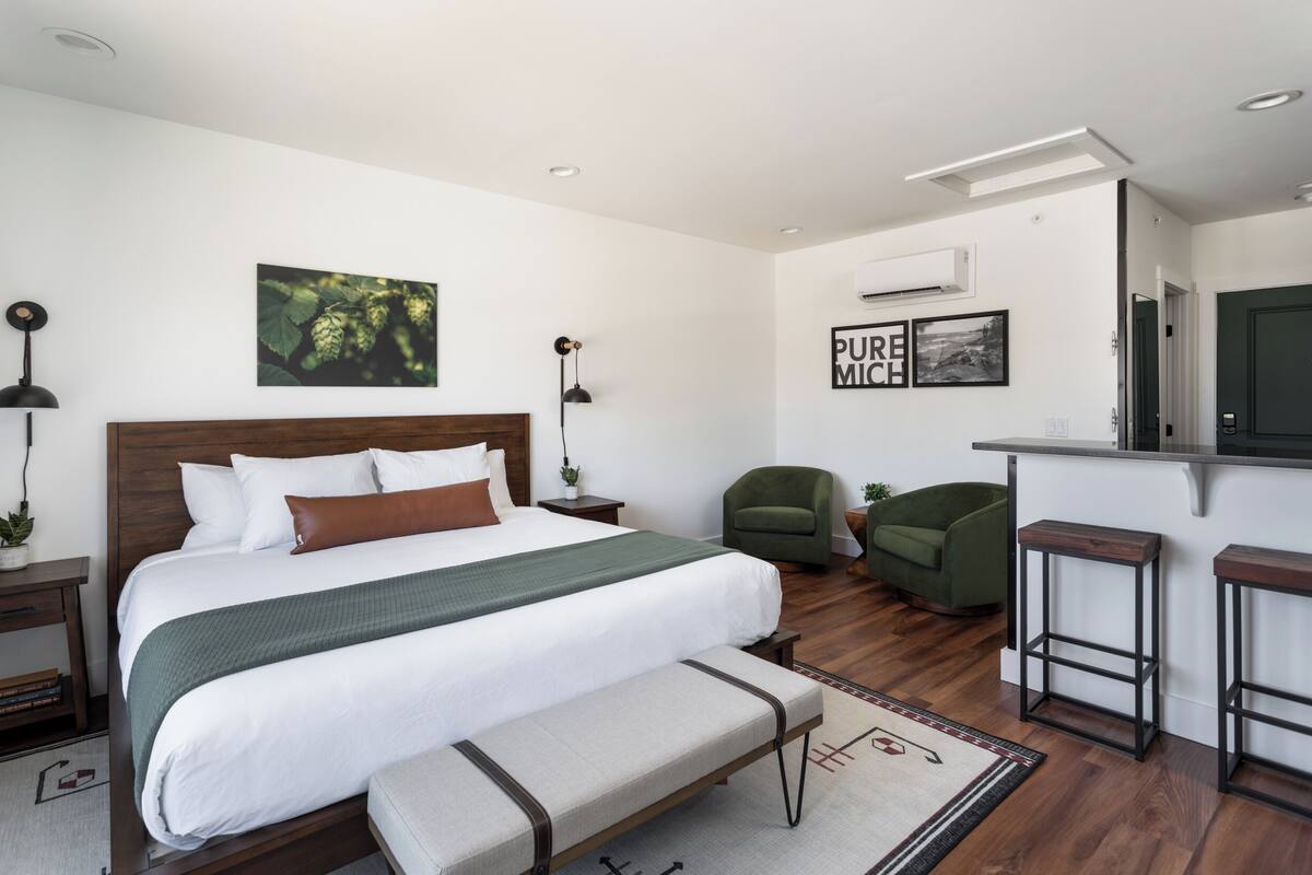 A modern hotel room with a large bed, a bench, a desk with stools, a green armchair, and artwork on the walls.