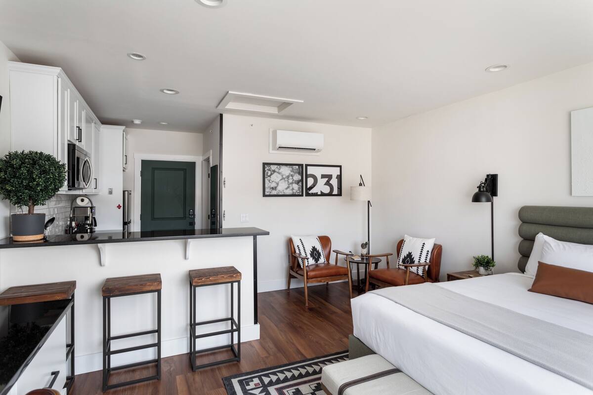 A modern hotel room with a kitchenette, a bed, a seating area with chairs, and a small table.