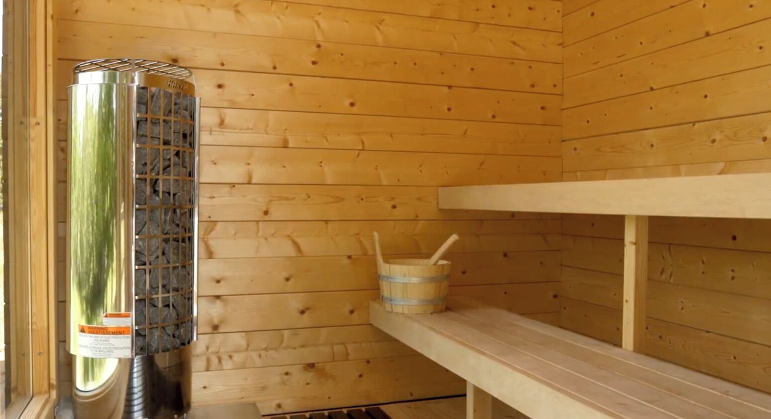 Una sauna de madera con un calentador metálico, un banco de madera y un cubo con una cuchara.