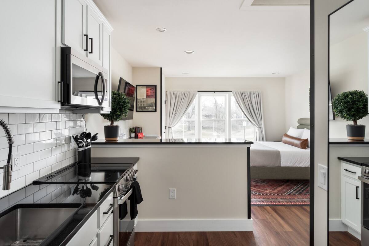A compact kitchen with white cabinets, a stove, and a microwave. There is a small island with a sink and a potted plant. A bed is visible in the background with a window and curtains.