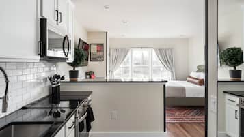 A compact kitchen with white cabinets, a stove, and a microwave. There is a small island with a sink and a potted plant. A bed is visible in the background with a window and curtains.