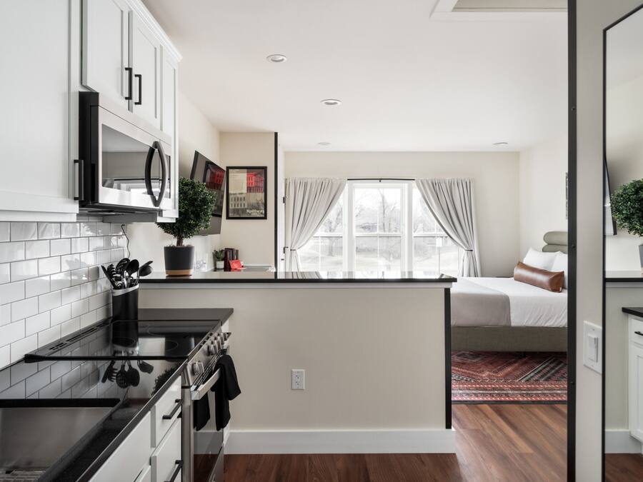 A compact kitchen with white cabinets, a stove, and a microwave. There is a small island with a sink and a potted plant. A bed is visible in the background with a window and curtains.