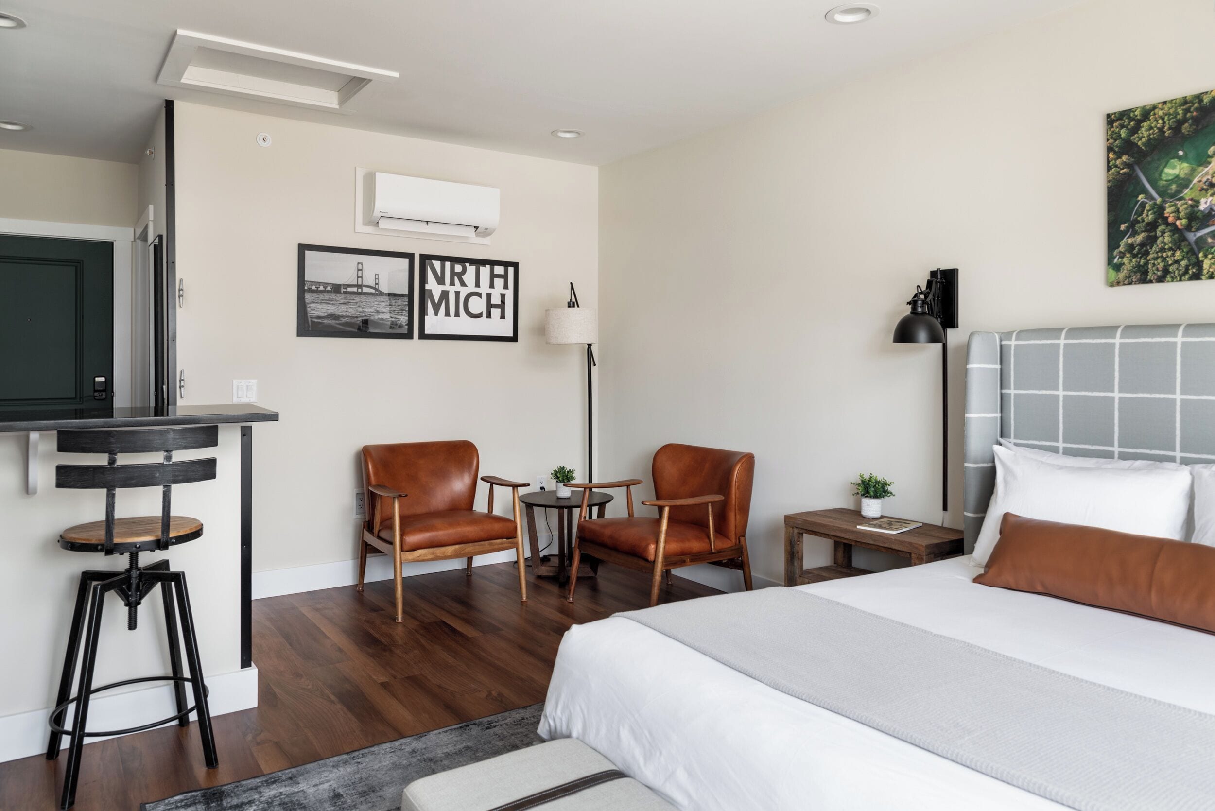 A modern hotel room with a bed, two leather chairs, a small table, and a desk with a stool.