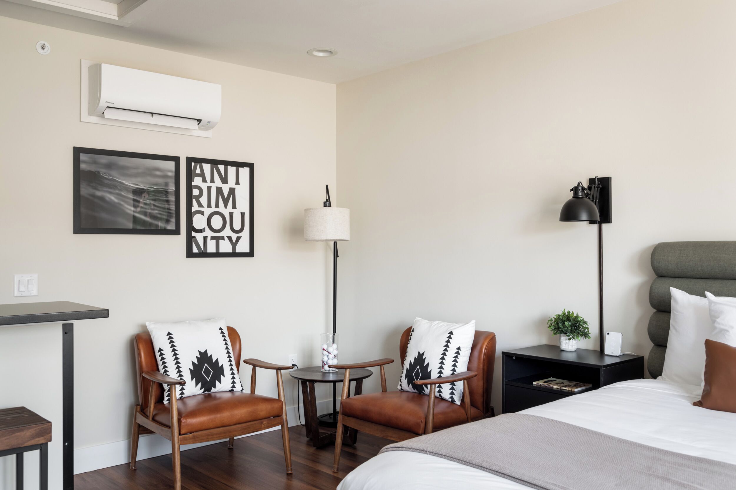 A modern hotel room with a bed, two leather chairs, a small table, a lamp, and a wall-mounted air conditioner.