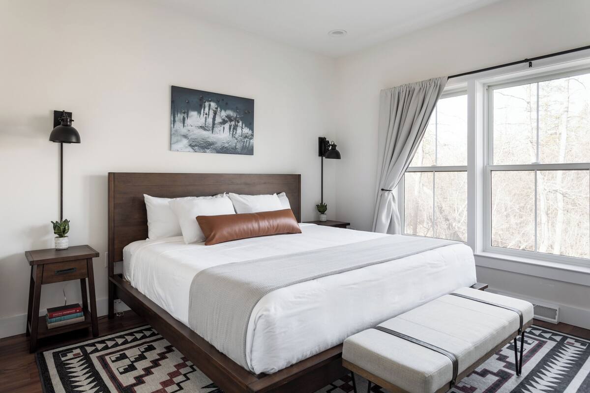 A neatly arranged bedroom with a large bed, a bedside table with a lamp, a bench, and a window with curtains.