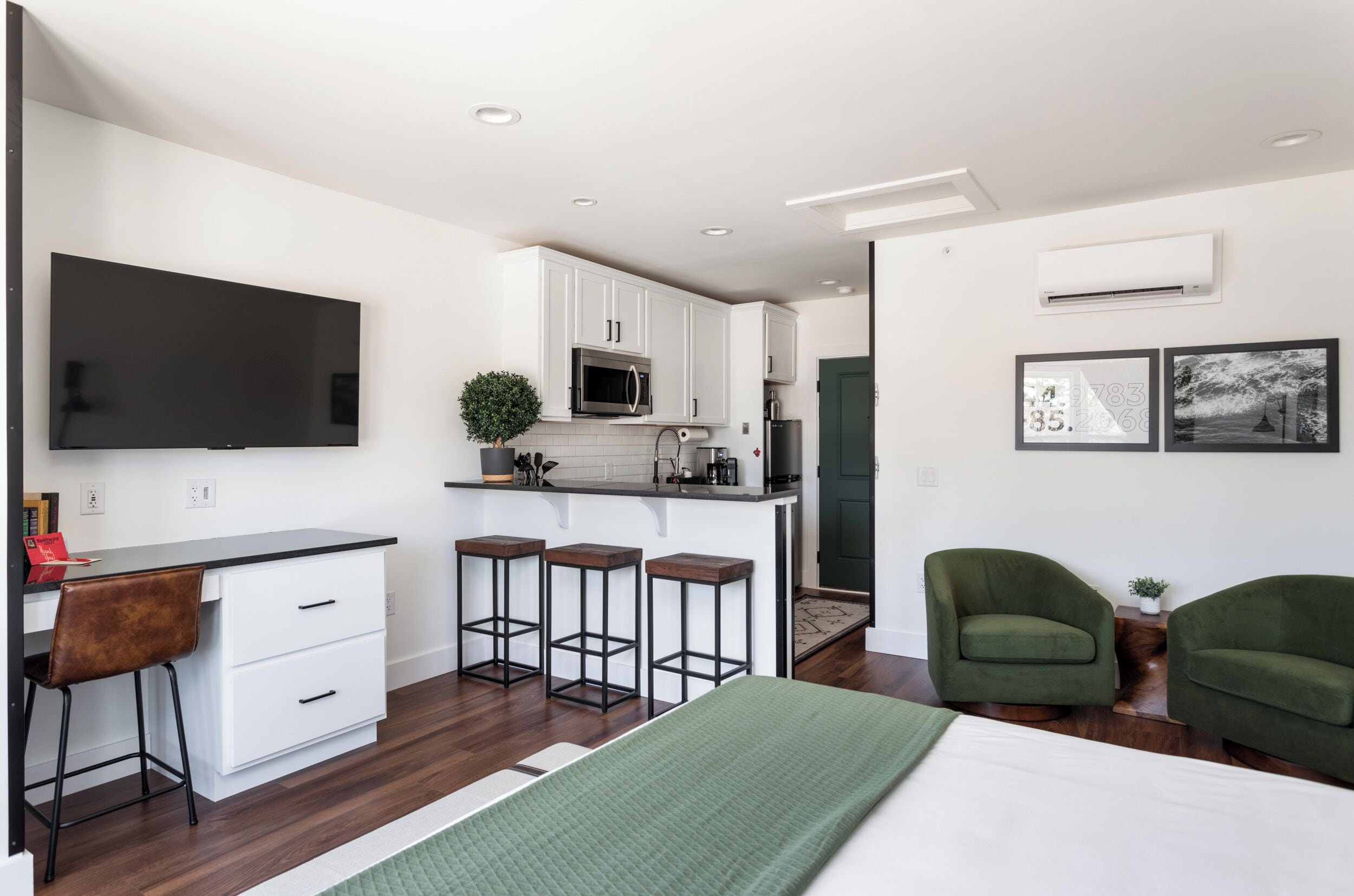 A modern kitchen with white cabinets, a microwave, and a large flat-screen TV. There are three bar stools and a green sofa in the living area.