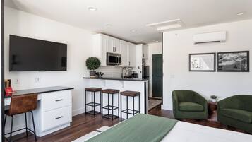 A modern kitchen with white cabinets, a microwave, and a large flat-screen TV. There are three bar stools and a green sofa in the living area.