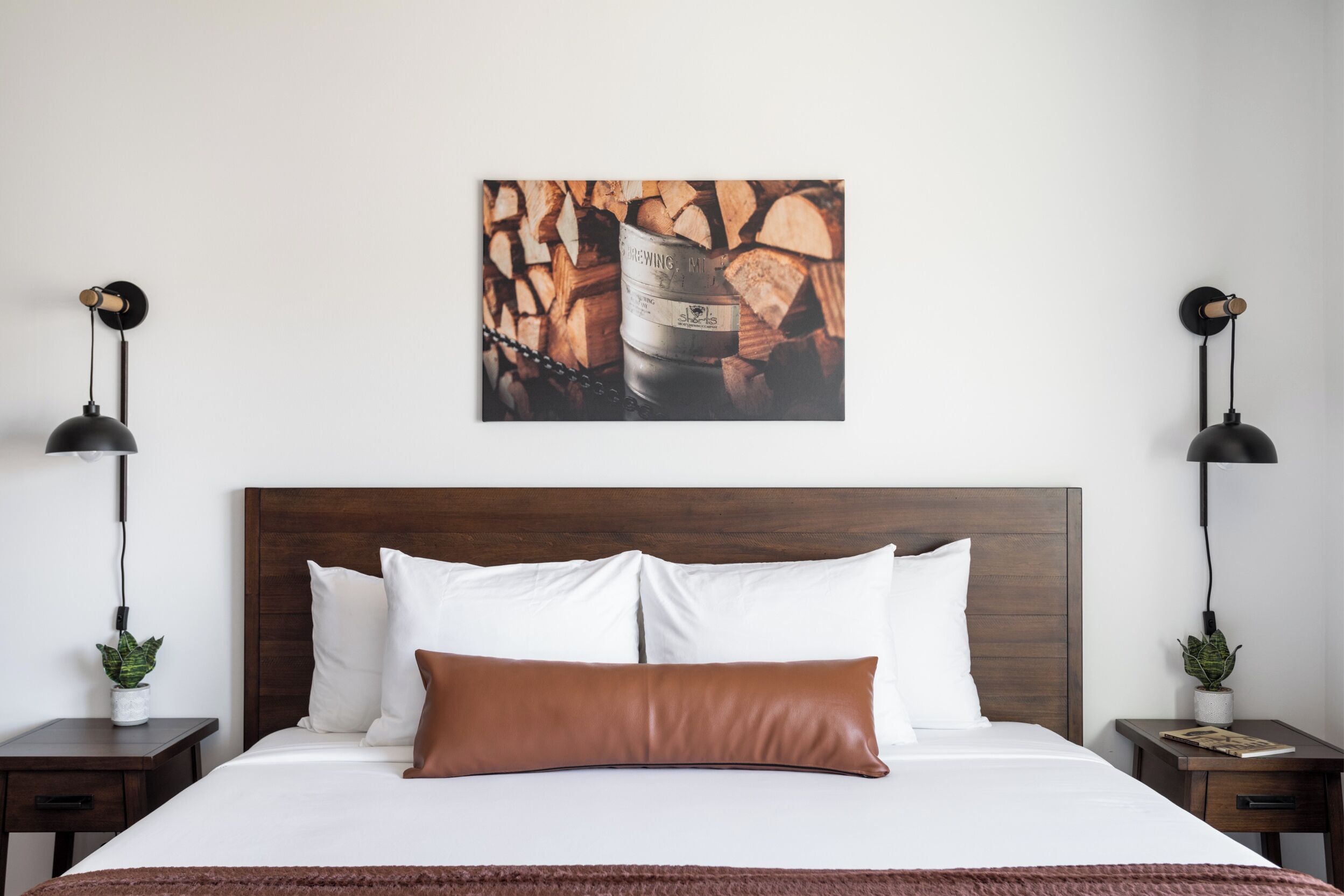 A neatly made bed with white and brown pillows, a wooden headboard, and a brown throw blanket. There are two bedside tables with lamps and small potted plants.