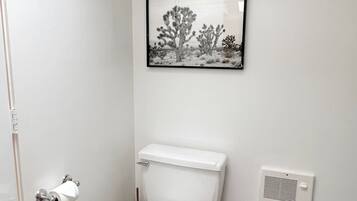 A bathroom with a toilet, a framed picture of a desert landscape, a trash bin, and a vent.