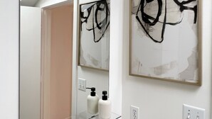 A bathroom with a large mirror, a glass shelf, a sink, and two abstract paintings on the wall.