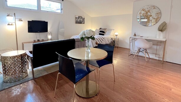 A modern, well-lit room with a bed, a dining area with a table and chairs, a TV mounted on the wall, and a large window providing natural light.