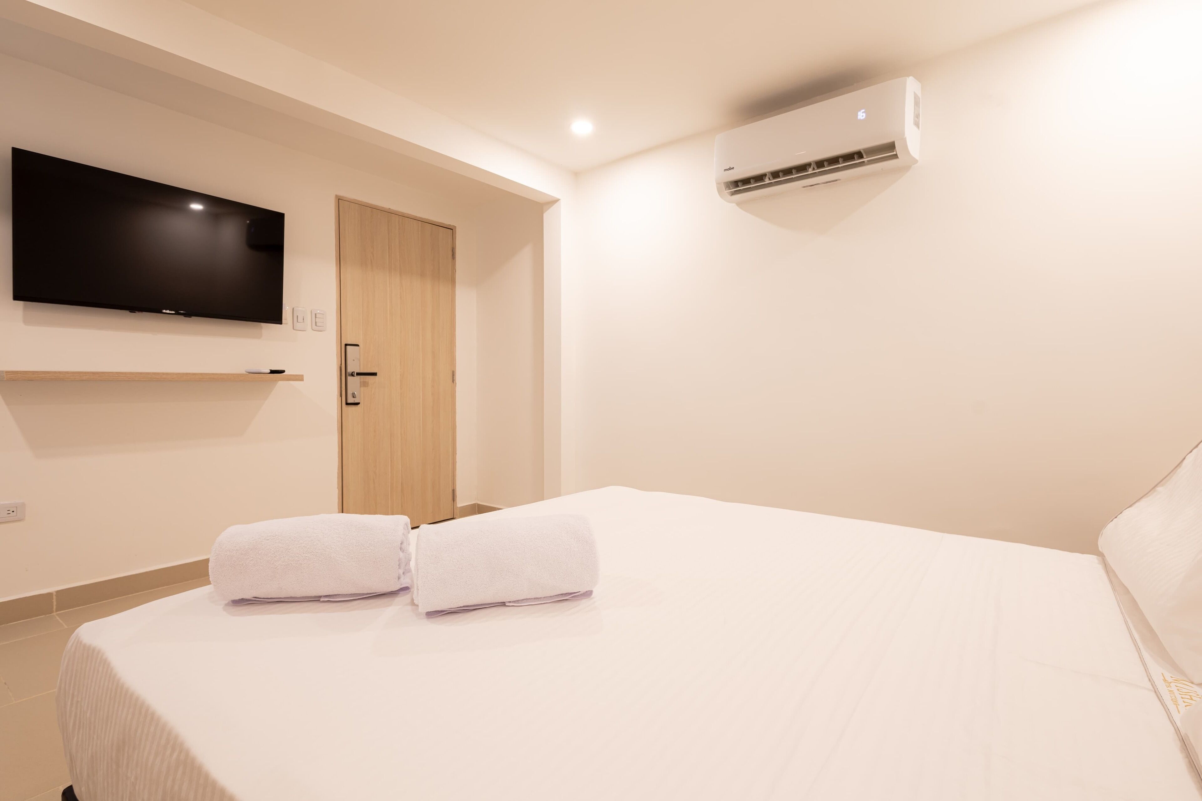 A hotel room with a bed, a flat-screen TV mounted on the wall, an air conditioning unit, and a wooden door.
