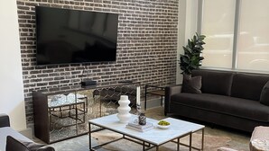 A modern living room with a brick wall, a flat-screen TV, a coffee table, a sofa, and a large window. - Sleek & Sophisticated: Modern Luxe in the Heart of New Orleans (New Orleans)
