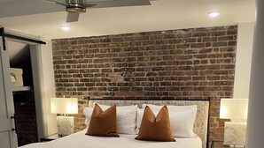 A modern bedroom with a brick wall, a large bed with white bedding and brown pillows, two bedside lamps, and a ceiling fan. - Sleek & Sophisticated: Modern Luxe in the Heart of New Orleans (New Orleans)