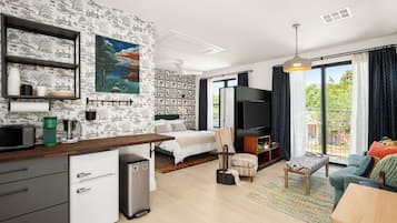 A modern hotel room with a kitchenette, a bed, a TV, a balcony with curtains, and a mural on the wall.