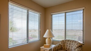 A room with two large windows with blinds, a small table with a lamp, and a patterned armchair. - Newly Remodeled Beautiful Home and Large Sparkling Pool, New Hot Tub & Game Room (San Tan Valley)